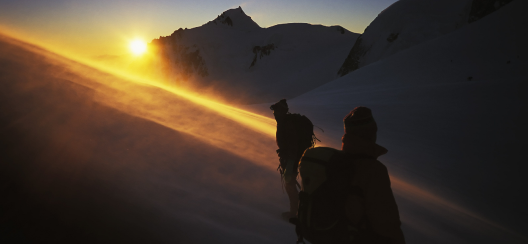 Two individuals stand on a mountain's edge, silhouetted against a vibrant sunset in the background.