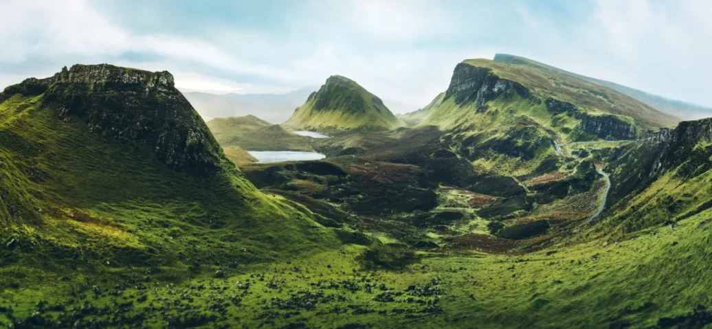 Lush green valley with rugged hills and a small lake under a cloudy sky.