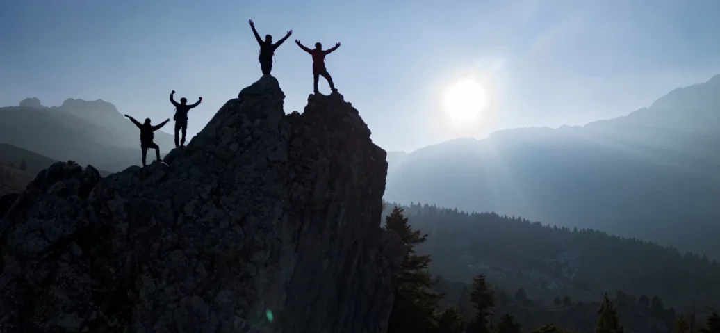 Individuals atop a mountain raise their arms in joy, celebrating their achievement with a stunning panoramic view.