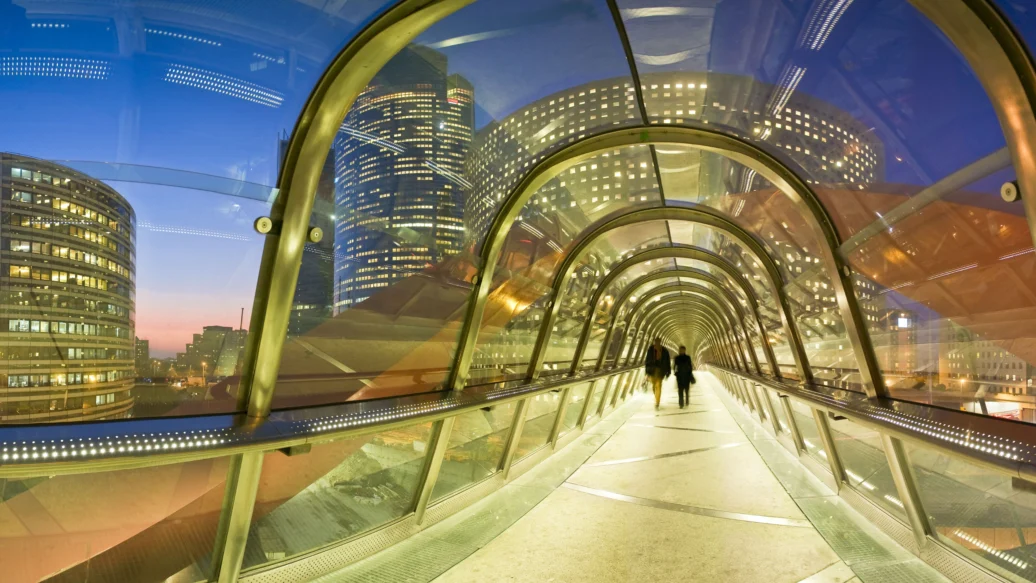 Glass-enclosed footbridge at dusk with city lights and a walking silhouette.