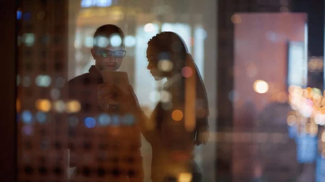 A man and woman stand together in front of a window, illuminated by soft night light, creating a serene atmosphere.