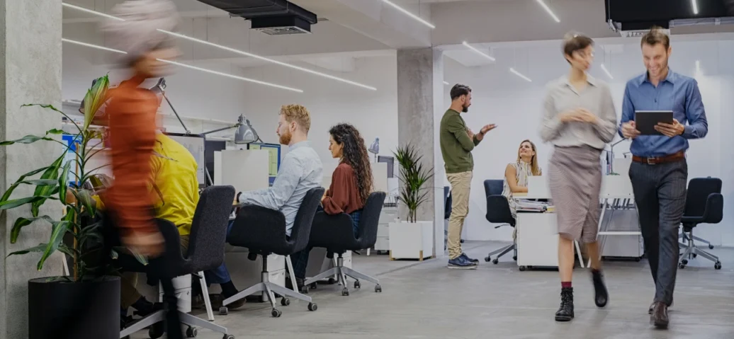 Office scene featuring people walking among desks and chairs, illustrating a busy and collaborative work atmosphere.