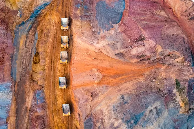 Aerial view of a dirt path with six dump trucks surrounded by colorful mineral terrain.