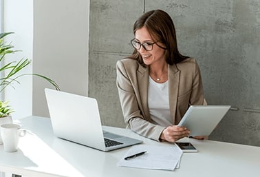 Woman with glasses looking at laptop while holding a tablet