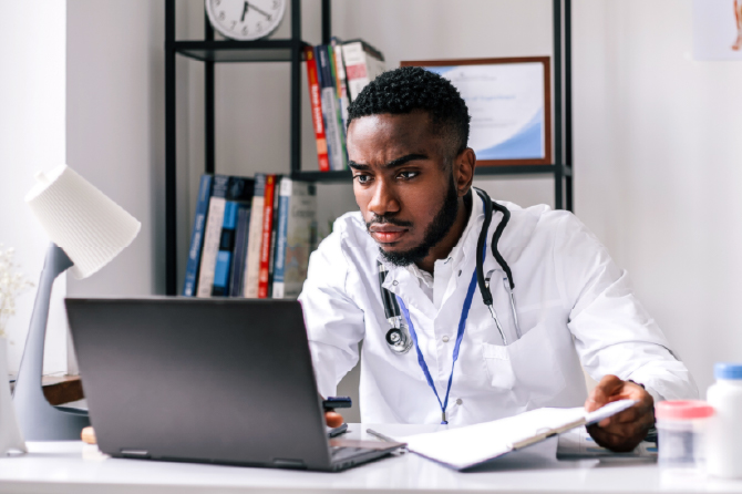 A person in a lab coat with a stethoscope works on a laptop, with books and a certificate in the background