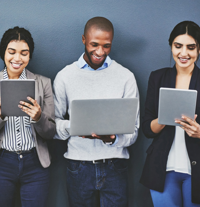 Three people smiling while using electronic devices against a gray wall